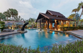 A pool with a waterfall element surrounded by chairs, umbrellas, and palm trees at The Slate, a Phuket Pearl Resort