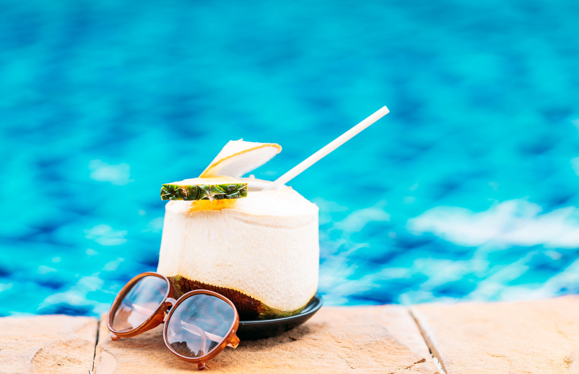 A coconut juice drink and a pair of sunglasses sit by the edge of a pool.