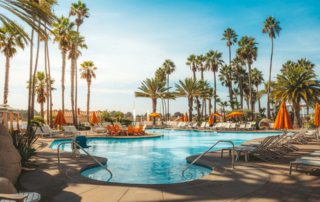 A hotel pool is surrounded by palm trees and orange umbrellas.