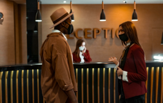 A guest and a hotel staff member wearing masks, talk at the reception desk.