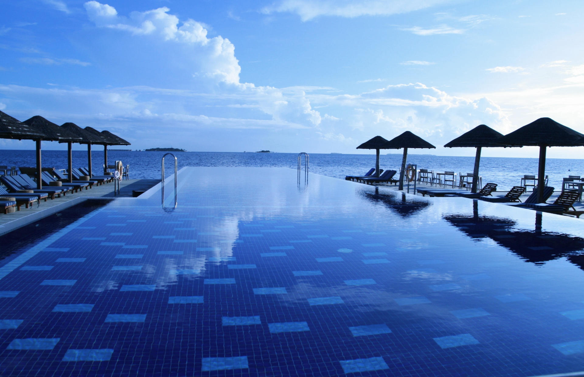 A hotel pool is lined with pool chairs and umbrellas and faces the ocean.