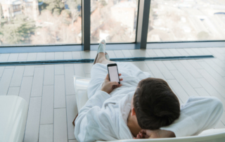 A man in a white robe reclining on a hotel chair looking at his phone.