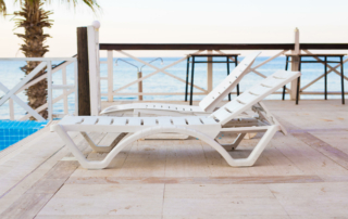 Two white beach chairs are positioned on a hotel pool deck by the ocean.