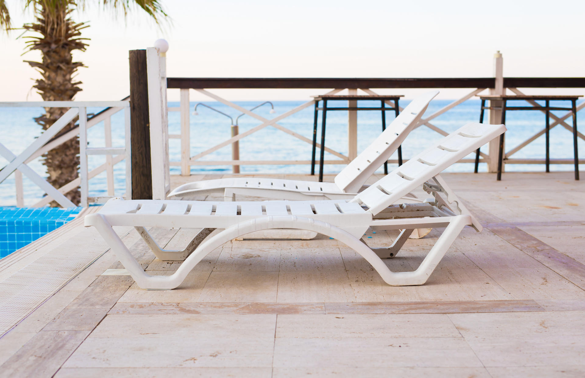 Two white beach chairs are positioned on a hotel pool deck by the ocean.