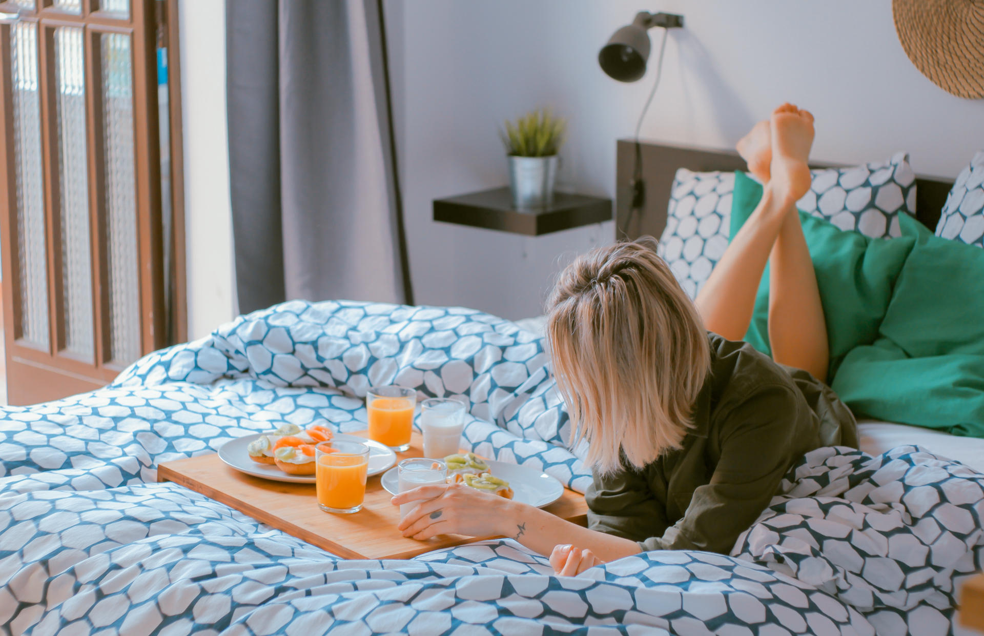 A woman is laying on her stomach on a hotel bed eating breakfast.