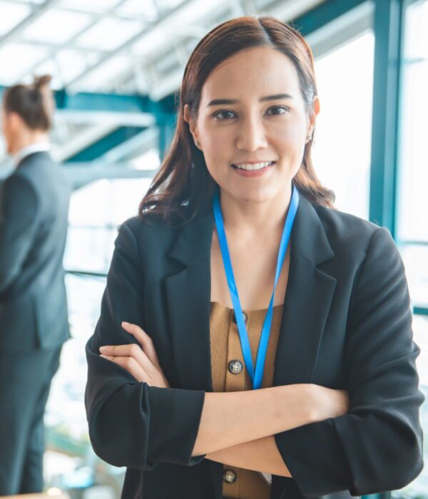 A business woman smiles at the camera with arms crossed while colleagues talk behind her.